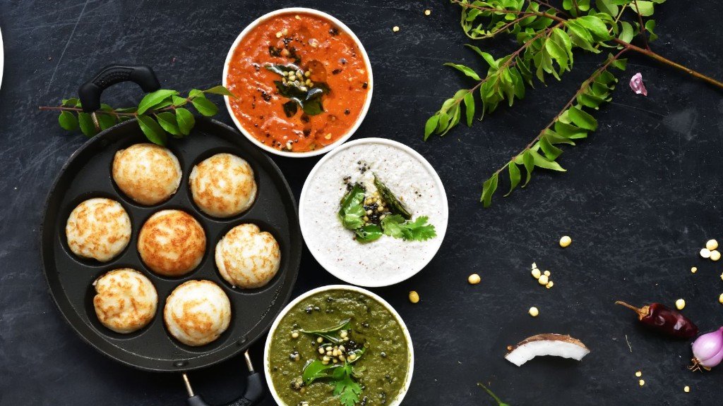 Golden kuzhi paniyaram in a traditional pan with tomato, coconut, and green chutneys